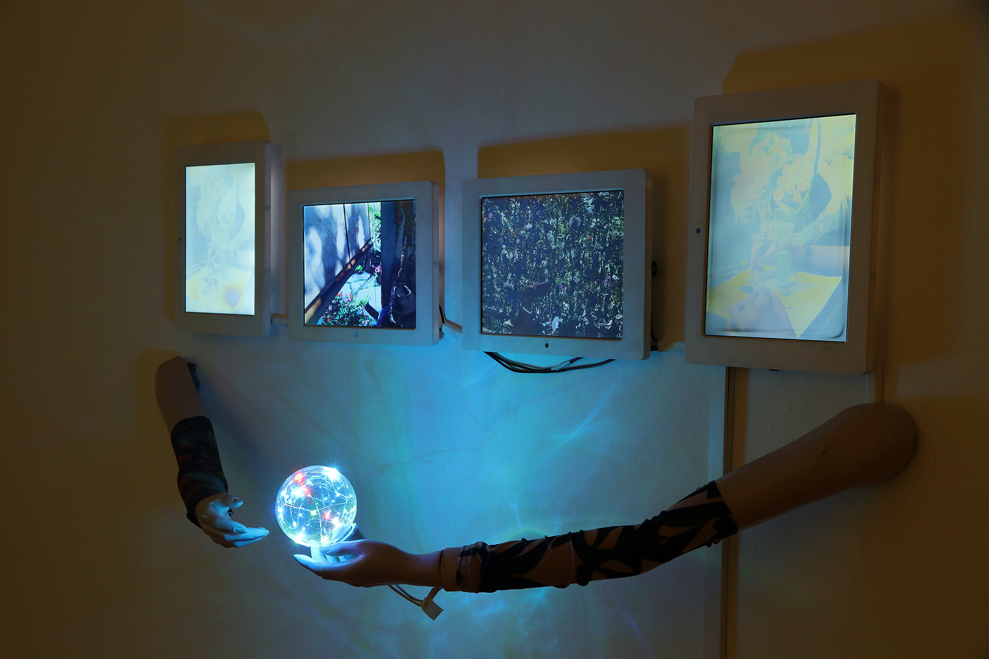 arms coming off a wall holding a glowing ball with ipads on the wall